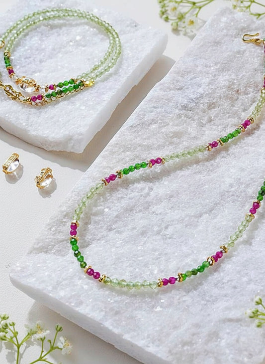 Beaded necklace with green, pink, and gold beads on a white surface with earrings and flowers.