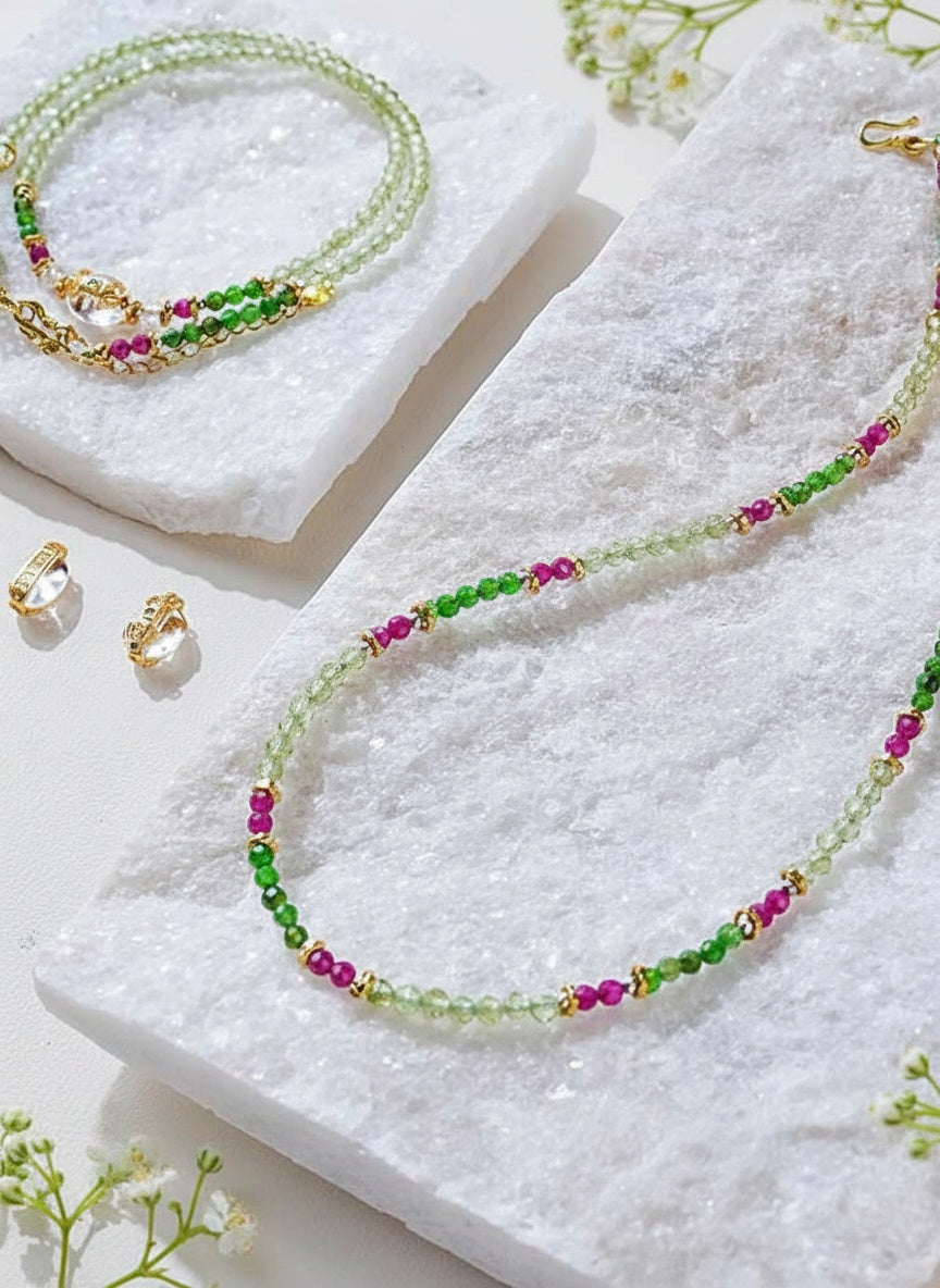 Beaded necklace with green, pink, and gold beads on a white surface with earrings and flowers.