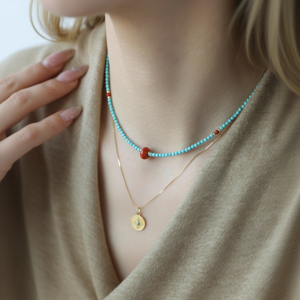 Necklace with turquoise beads and gold pendant worn by a person in a beige outfit.