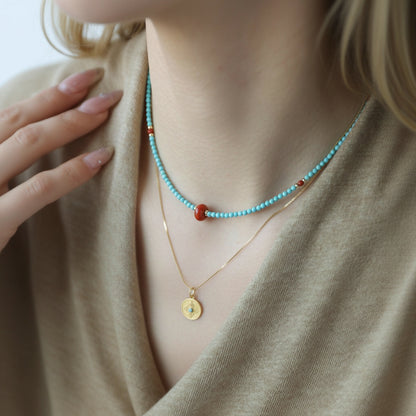 Necklace with turquoise beads and gold pendant worn by a person in a beige outfit.