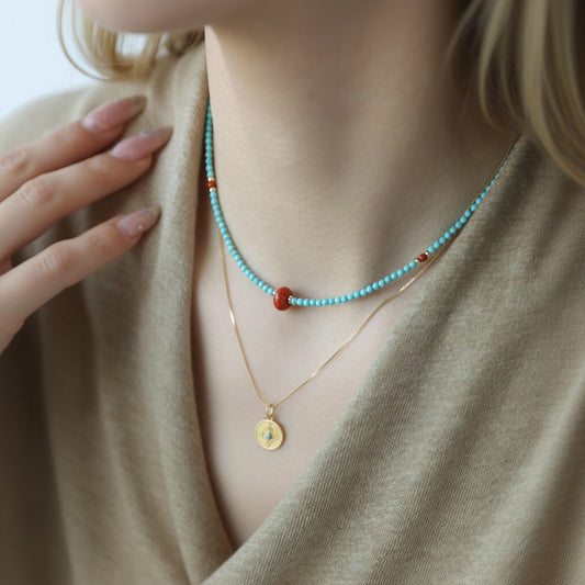 Necklace with turquoise beads and gold pendant worn by a person in a beige outfit.