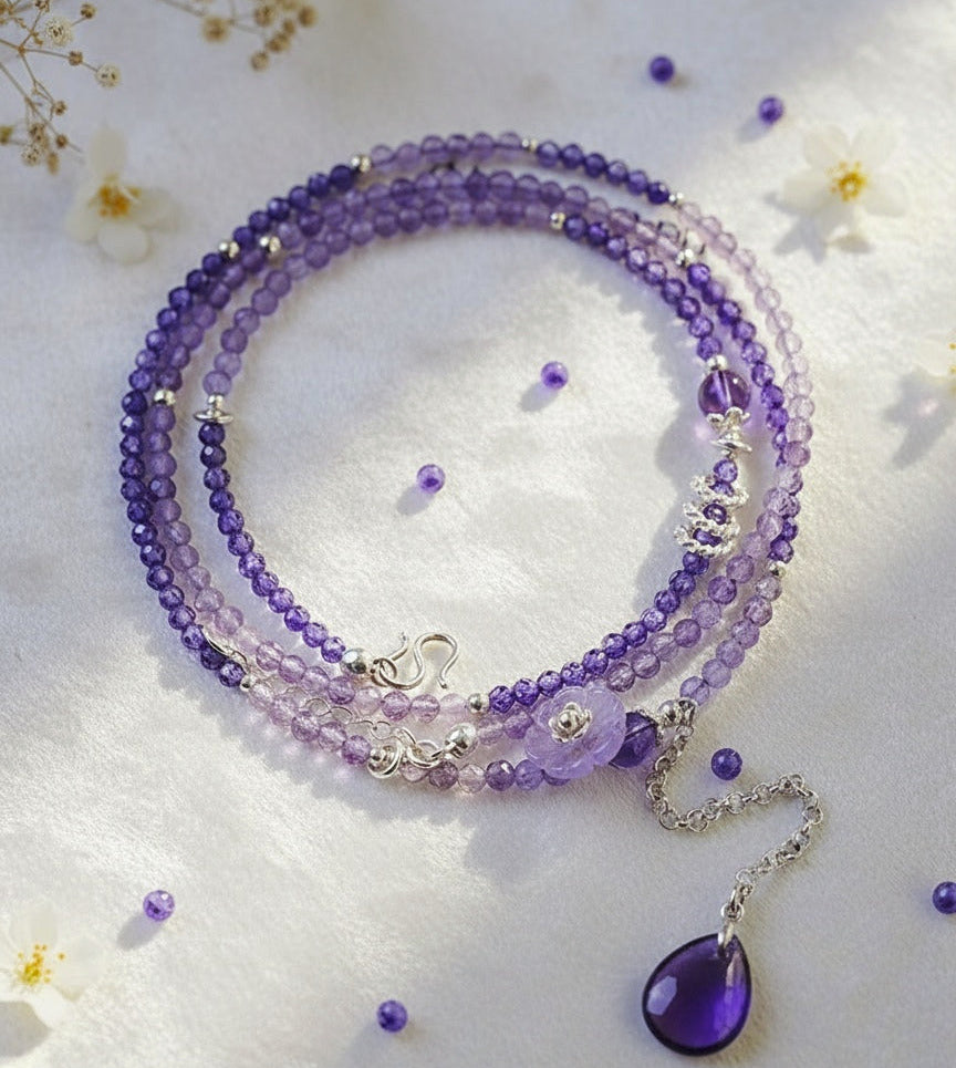 Purple beaded necklace with a teardrop pendant on a white background with small flowers.