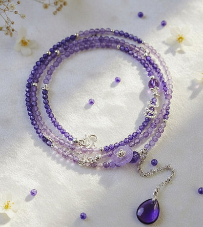 Purple beaded necklace with a teardrop pendant on a white background with small flowers.