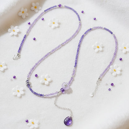 Purple beaded necklace with a teardrop pendant on a white surface with small flowers.