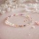 A multicolor tourmaline and freshwater pearl bracelet with gold-plated accents, displayed on a pink background with white flowers.