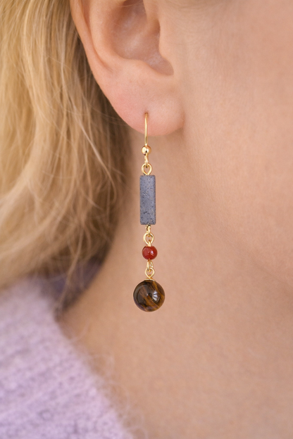 Earring with gold hook, blue stone, red bead, and brown round stone on a blurred background