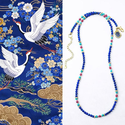 Beaded necklace with blue, red, and green beads on a white background, next to a decorative fabric with cranes and flowers.