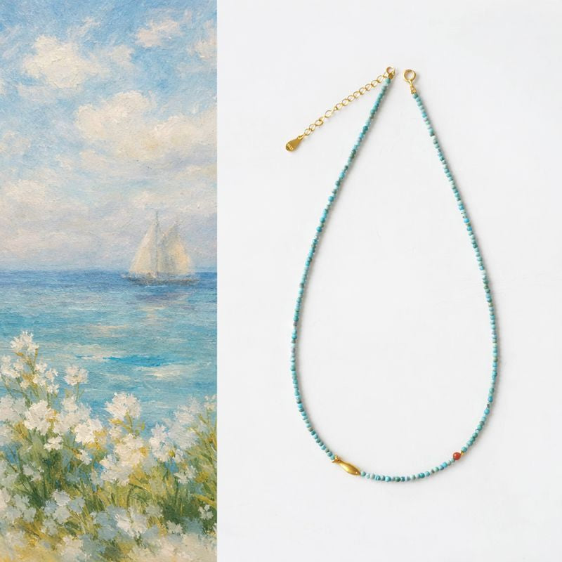 Necklace with blue beads and gold clasp on a white background, next to a painting of a sailboat on water.
