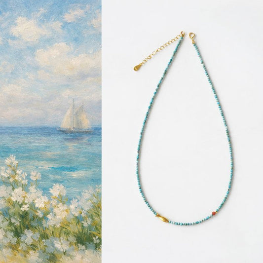 Necklace with blue beads and gold clasp on a white background, next to a painting of a sailboat on water.