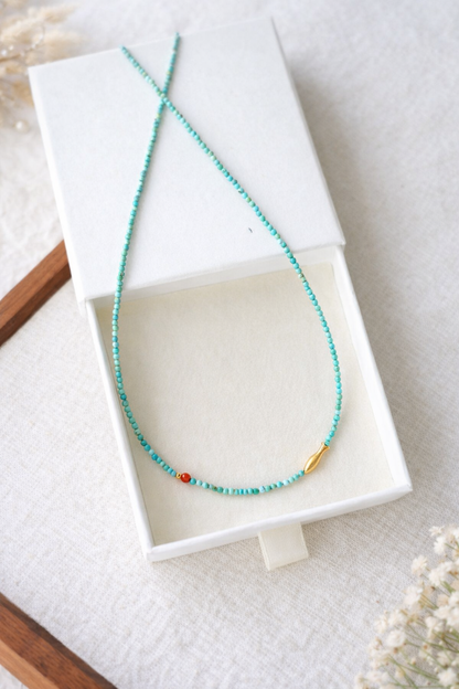 Turquoise beaded necklace with a red bead in a white jewelry box on a light background